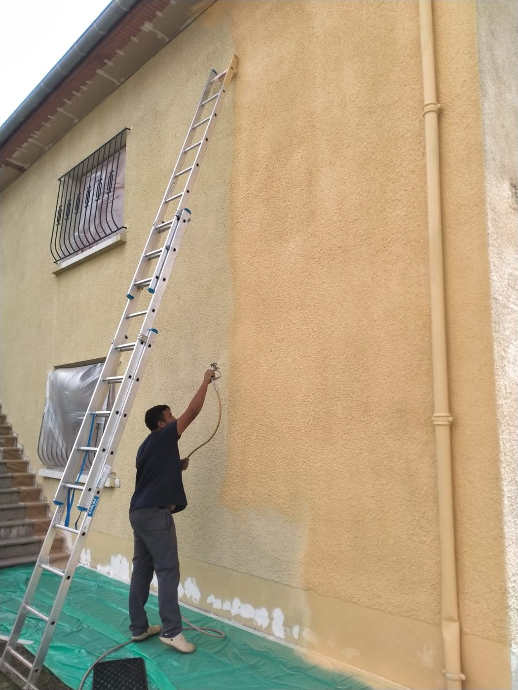 Artisan peintre de l'équipe Lambert appliquant une peinture de façade au pistolet, depuis une échelle